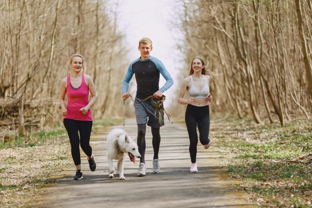 Three adults jogging with a dog on a forest path on a bright spring day, promoting healthy living.