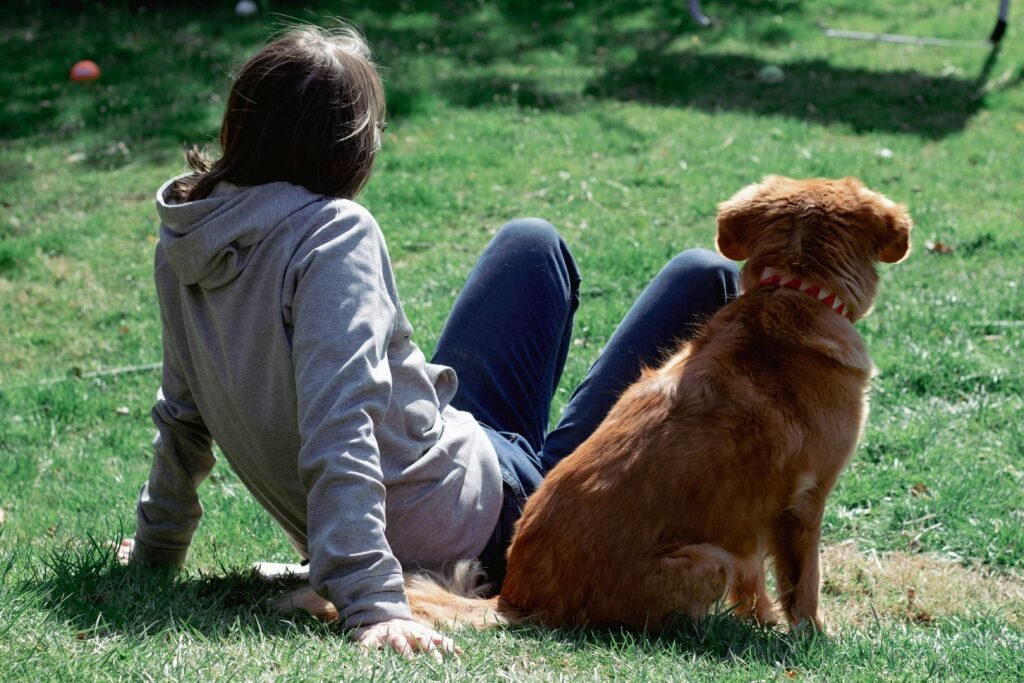 Woman and brown dog sitting together on green grass in a sunny park.